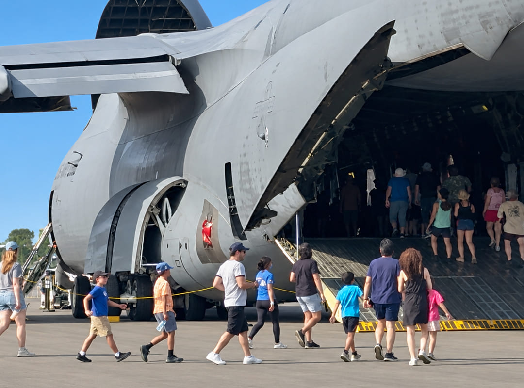 Air show visitors walk up the rear cargo ramp to explore the interior of a large U.S. military transport aircraft on static display.