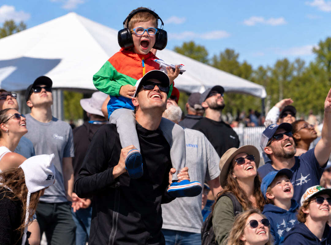 About A young child wearing hearing protection sits on an adult’s shoulders as spectators look skyward during an air show.