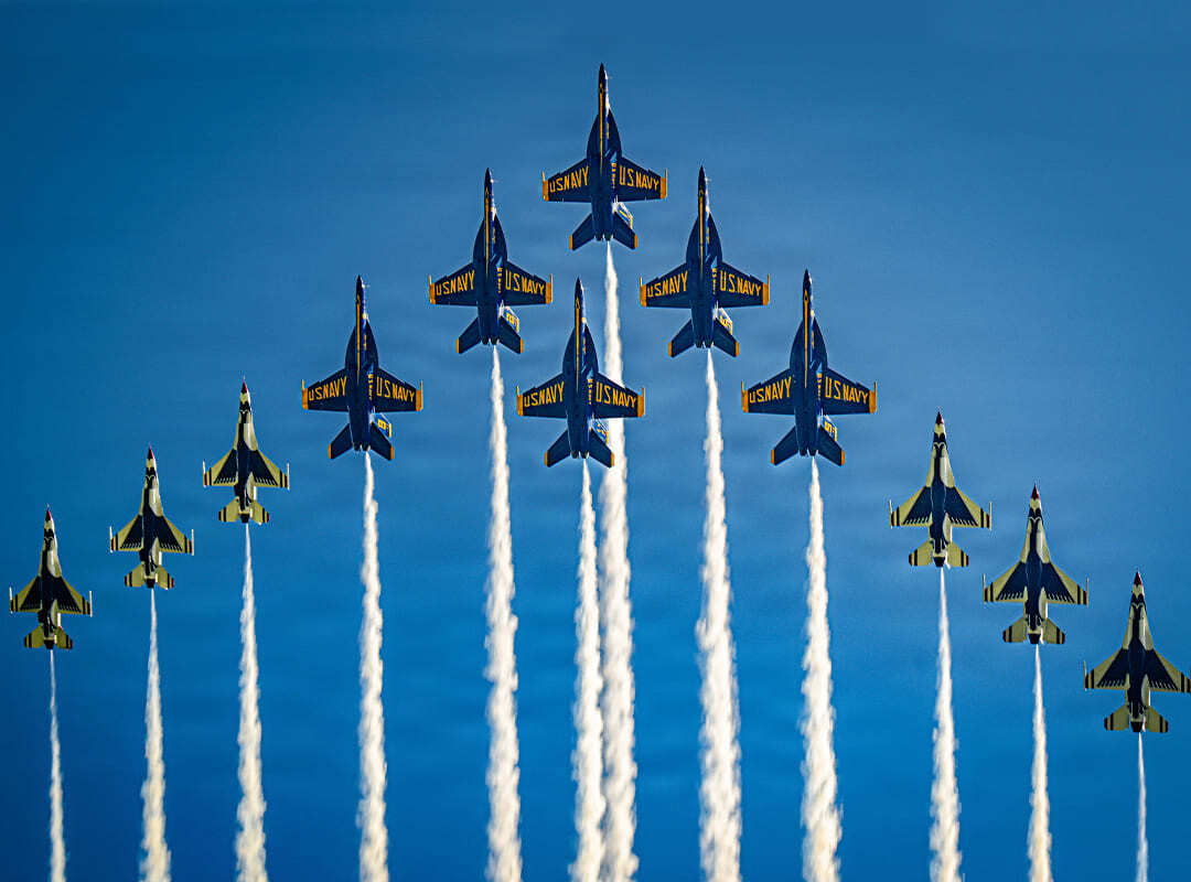 U.S. Navy Blue Angels and U.S. Air Force Thunderbirds fly in formation with smoke trails against a blue sky.