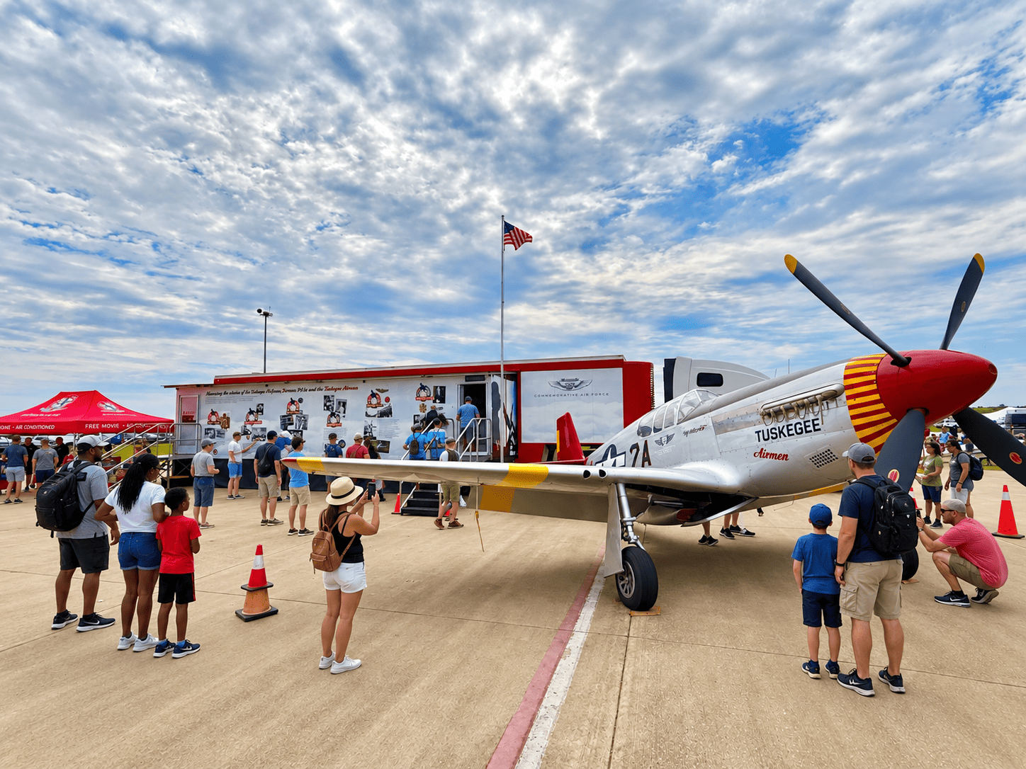Experiences Visitors gather around a P-51 Mustang honoring the Tuskegee Airmen, displayed beside a mobile aviation history exhibit at the air show.