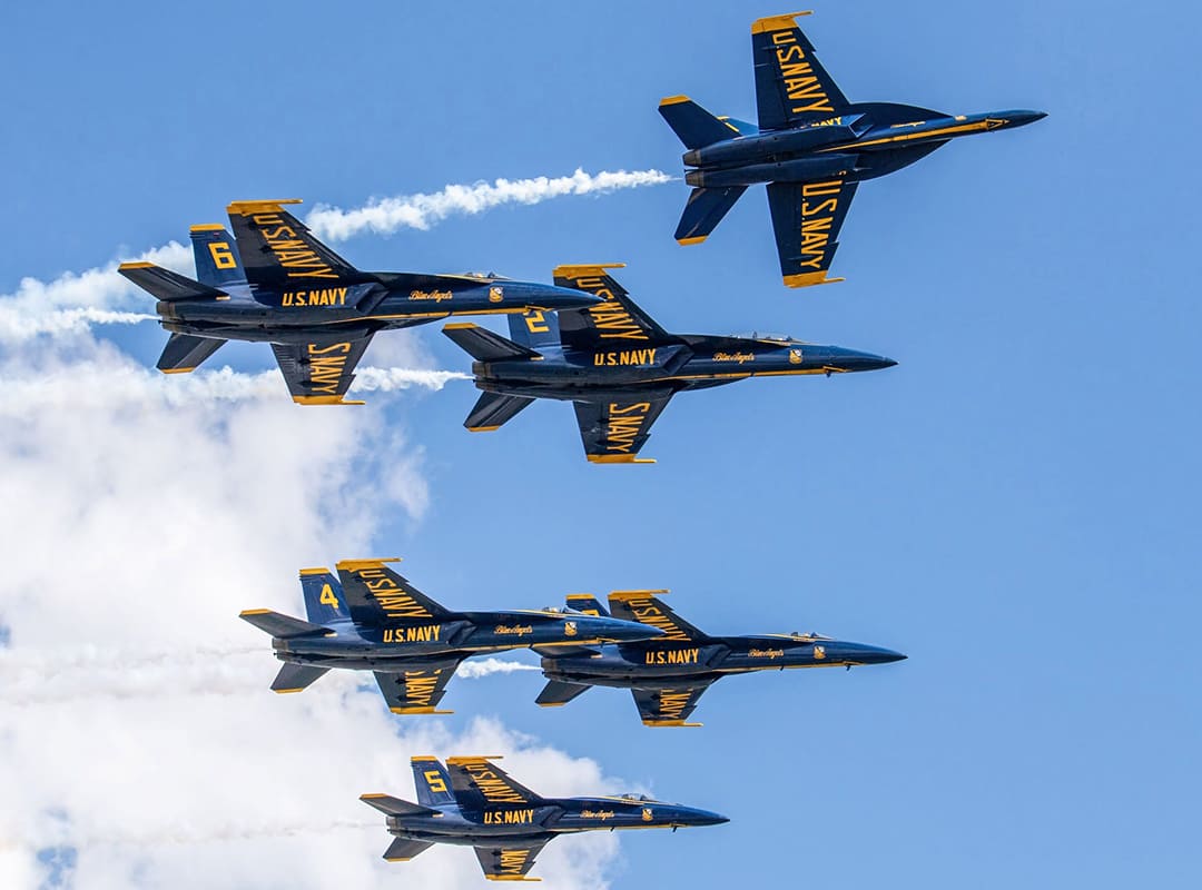 Six U.S. Navy Blue Angels F/A-18 Super Hornets climb in a tight formation, trailing white smoke against a clear blue sky.