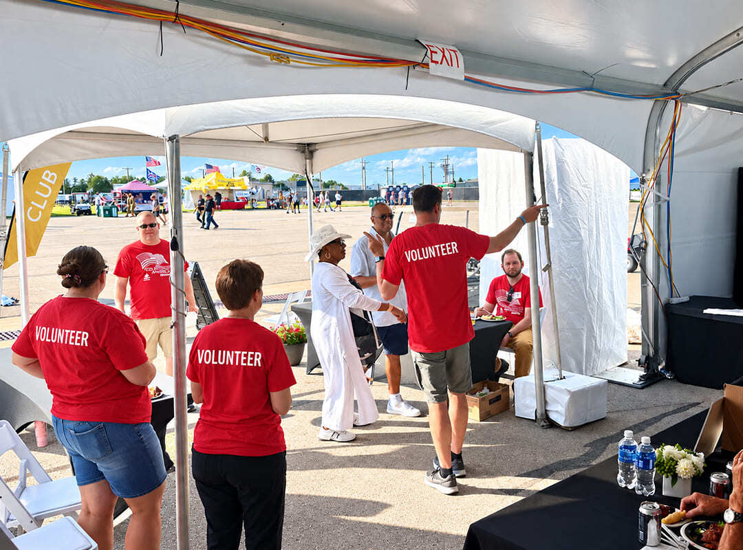 Volunteer Involvement Volunteers wearing red shirts assist guests beneath a hospitality tent at the airshow, with vendor booths and other event activities visible in the background